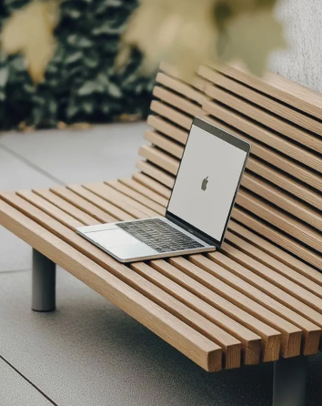 Open laptop with Apple logo on screen placed on a wooden slatted bench outdoors.