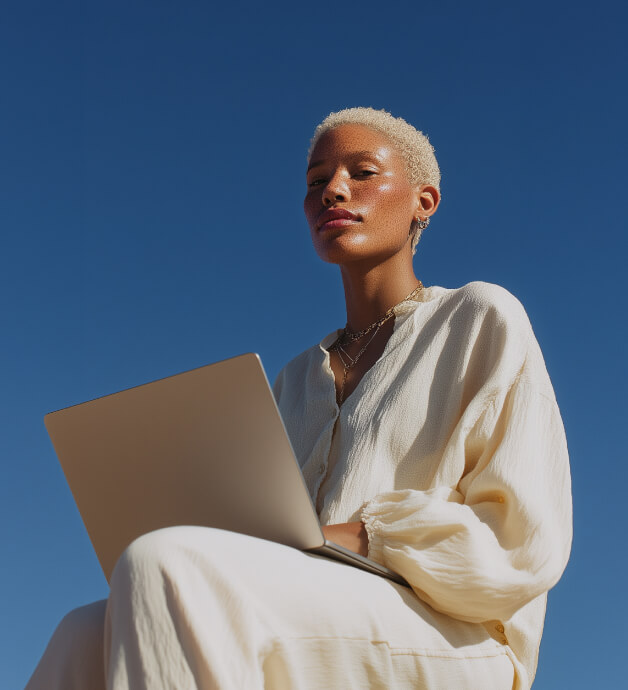 Young woman with short blonde hair sitting outdoors against a clear blue sky, working on a laptop while wearing a cream-colored blouse and pants.