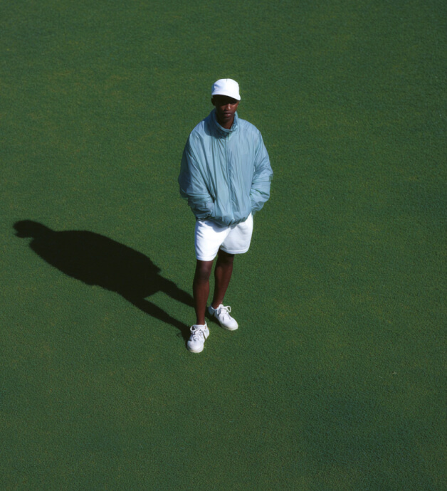 Man wearing a light blue jacket, white shorts, white cap, and white sneakers standing on green grass casting a shadow.