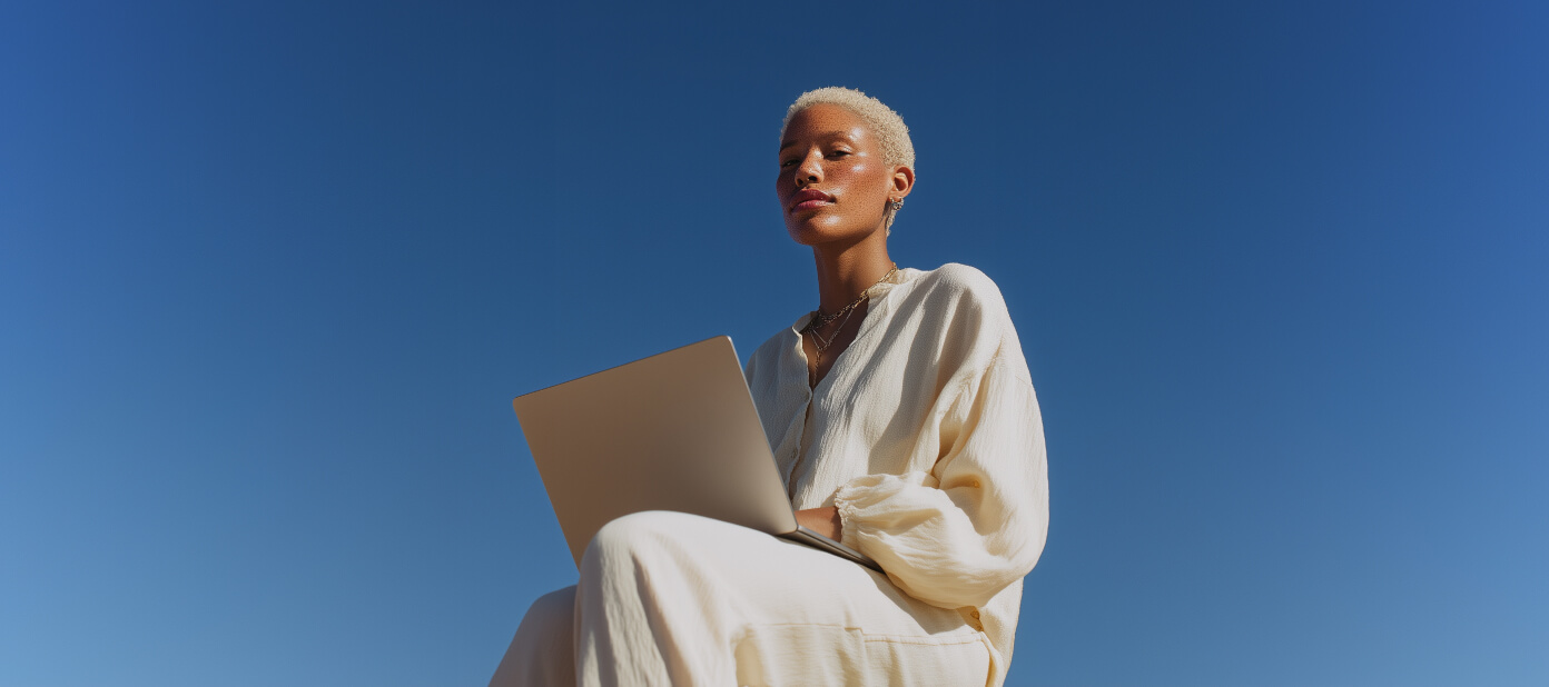 Person with short blonde hair wearing cream clothing sitting outdoors with a laptop against a clear blue sky.