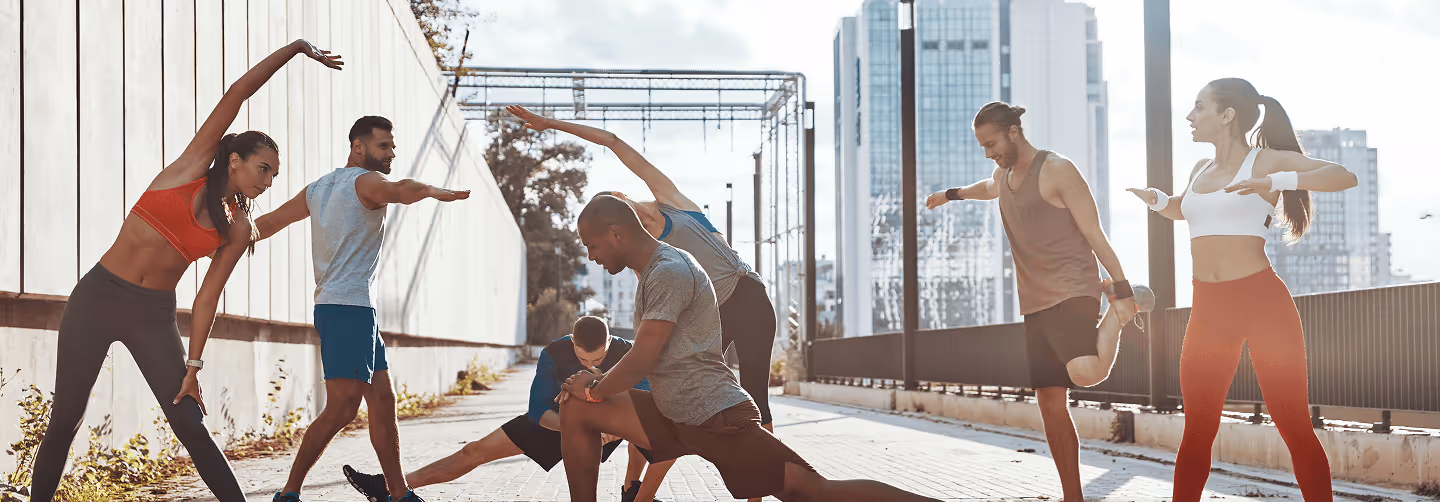 Group of six people stretching together outdoors in athletic wear on a sunny urban pathway.