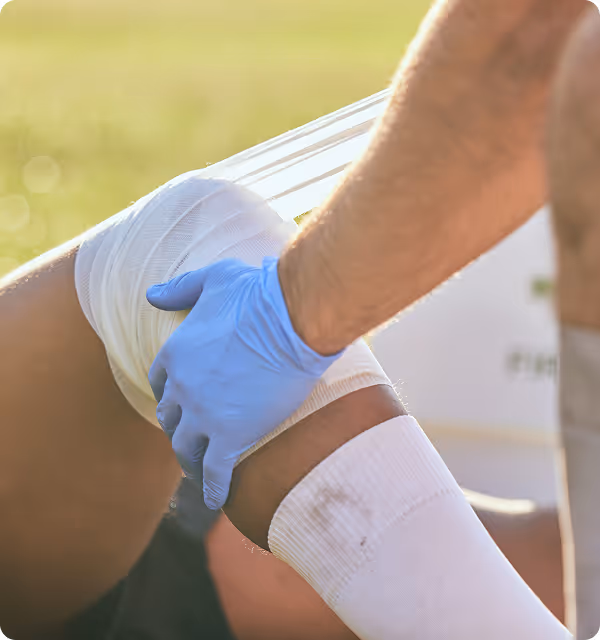 Person wearing blue gloves wraps a bandage around a knee outdoors in sunlight.