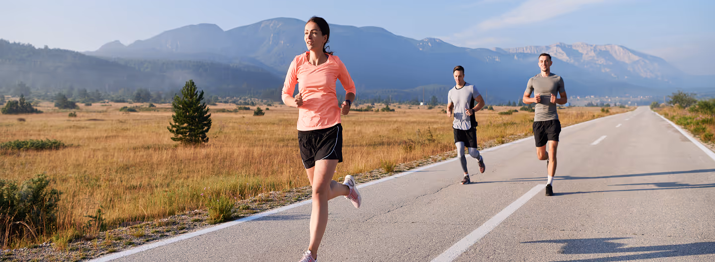 Three people jogging on a paved road in a rural area with mountains in the background during daytime.