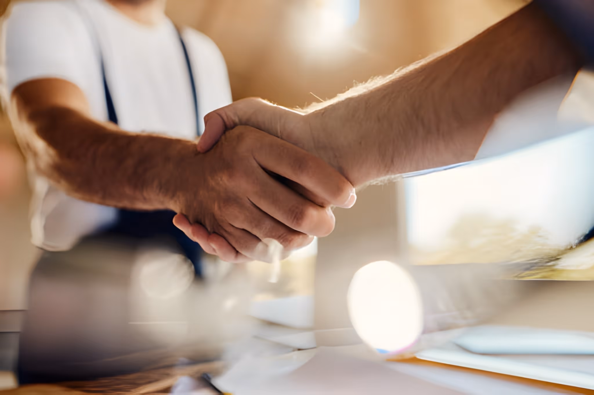 Close-up of two people shaking hands over a desk with documents and a laptop in soft focus.