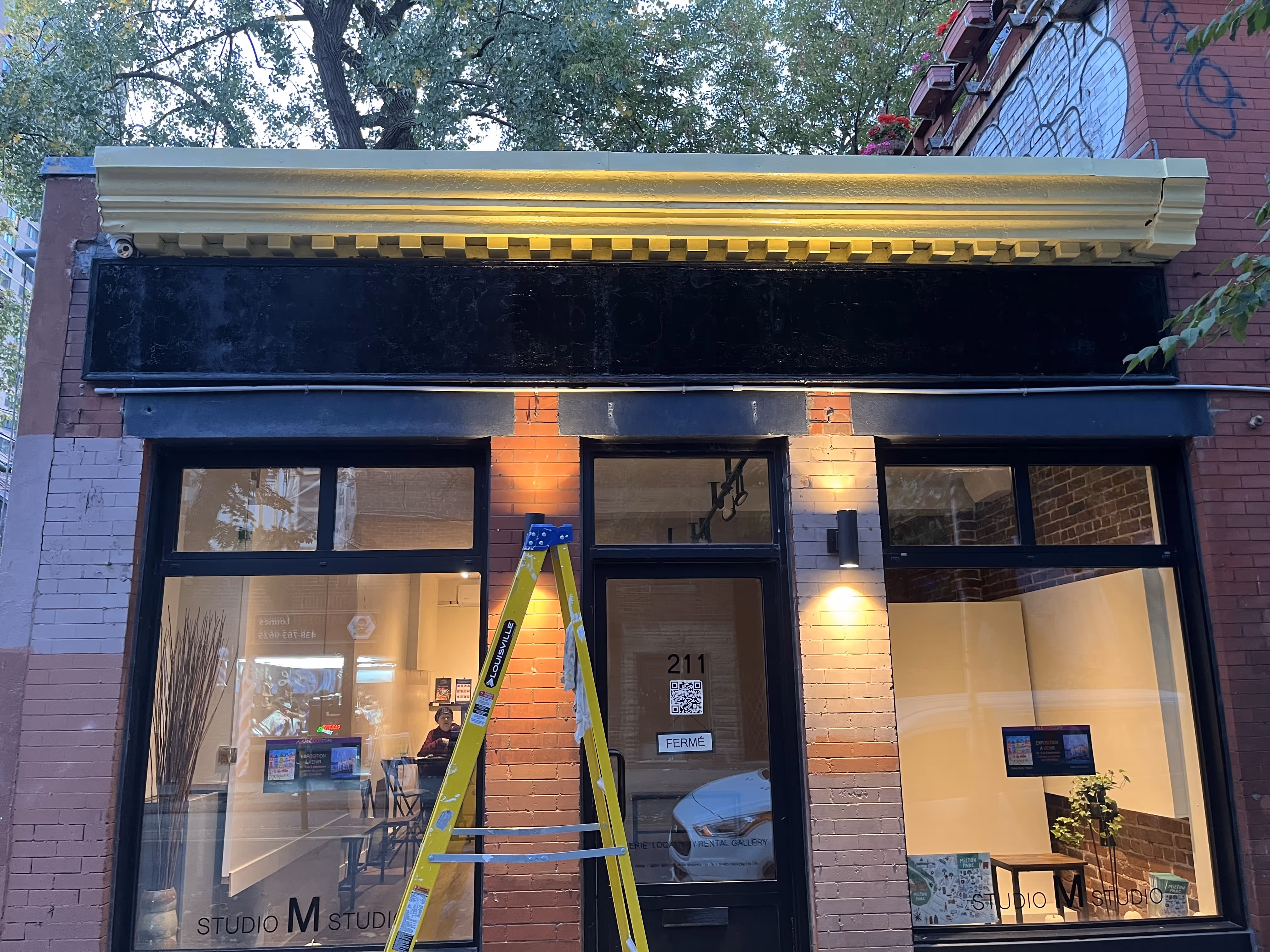 Front view of a small brick storefront with large windows, a closed black door labeled 211 with a 'FERMÉ' sign, yellow ladder leaning against the building, and interior lit with plants and chairs.