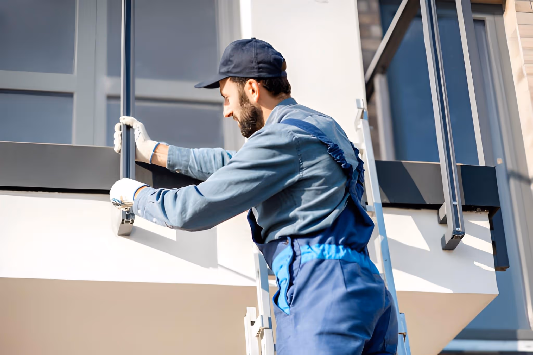 Worker in blue overalls and cap inspecting and cleaning a metal balcony railing outdoors.