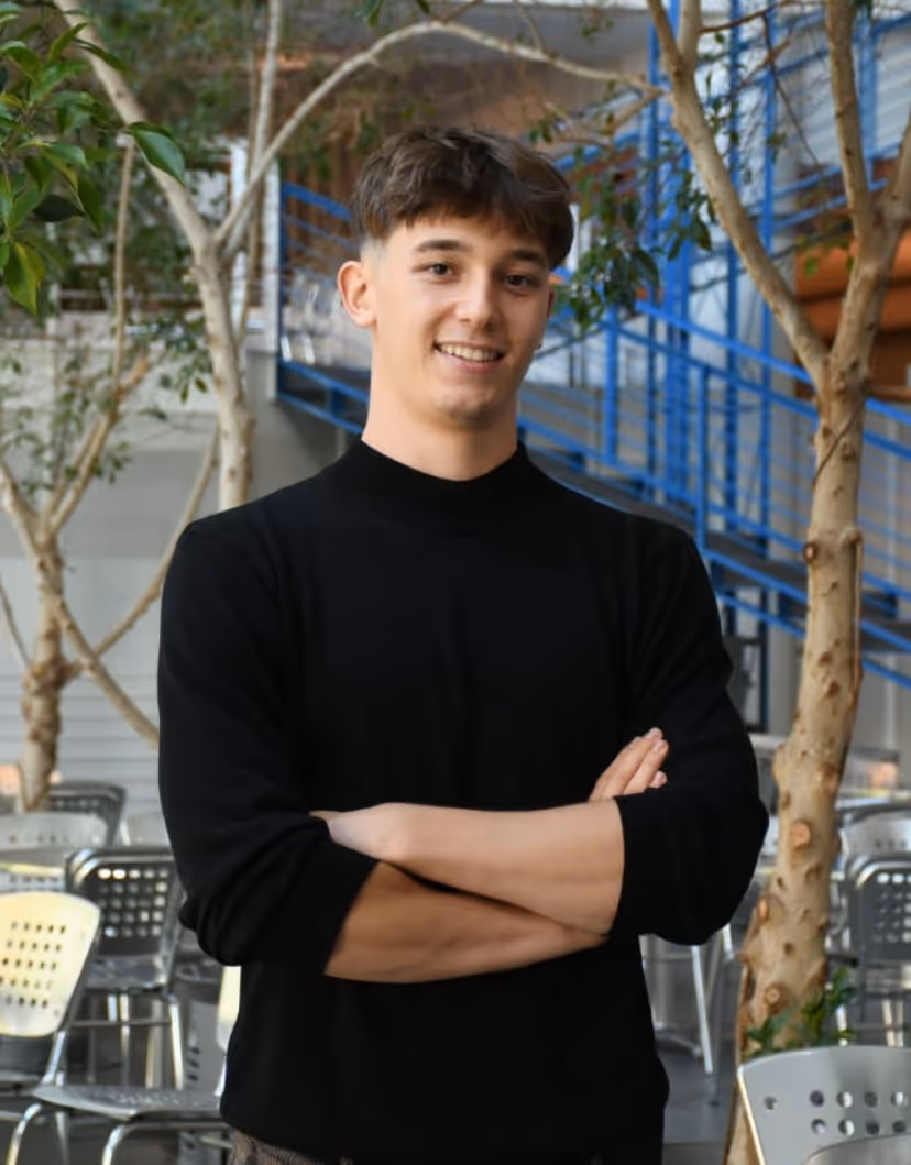 Young man with short brown hair smiling with arms crossed, standing in an indoor space with trees and chairs.