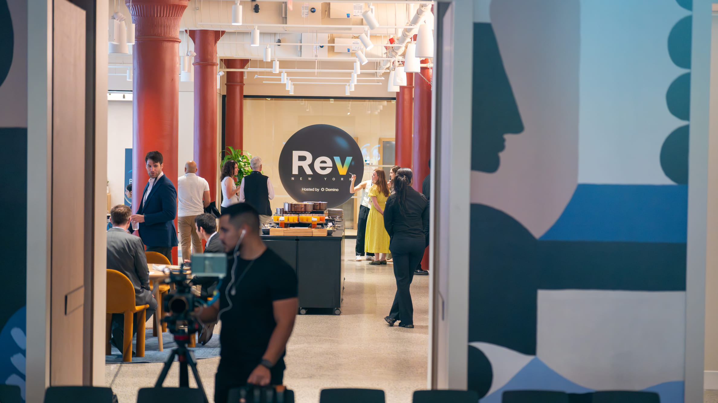 People networking and a cameraman filming at a Rev New York event with red pillars and artwork visible.