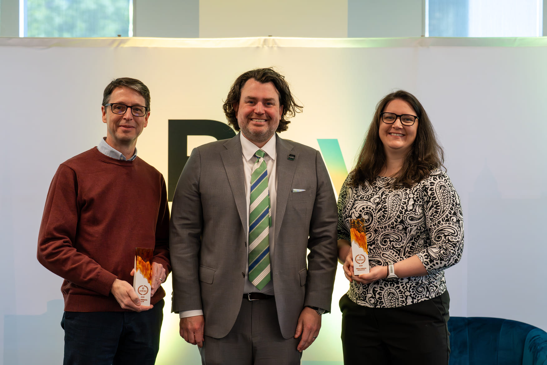 Two people holding awards standing beside a man in a gray suit with a striped green tie.