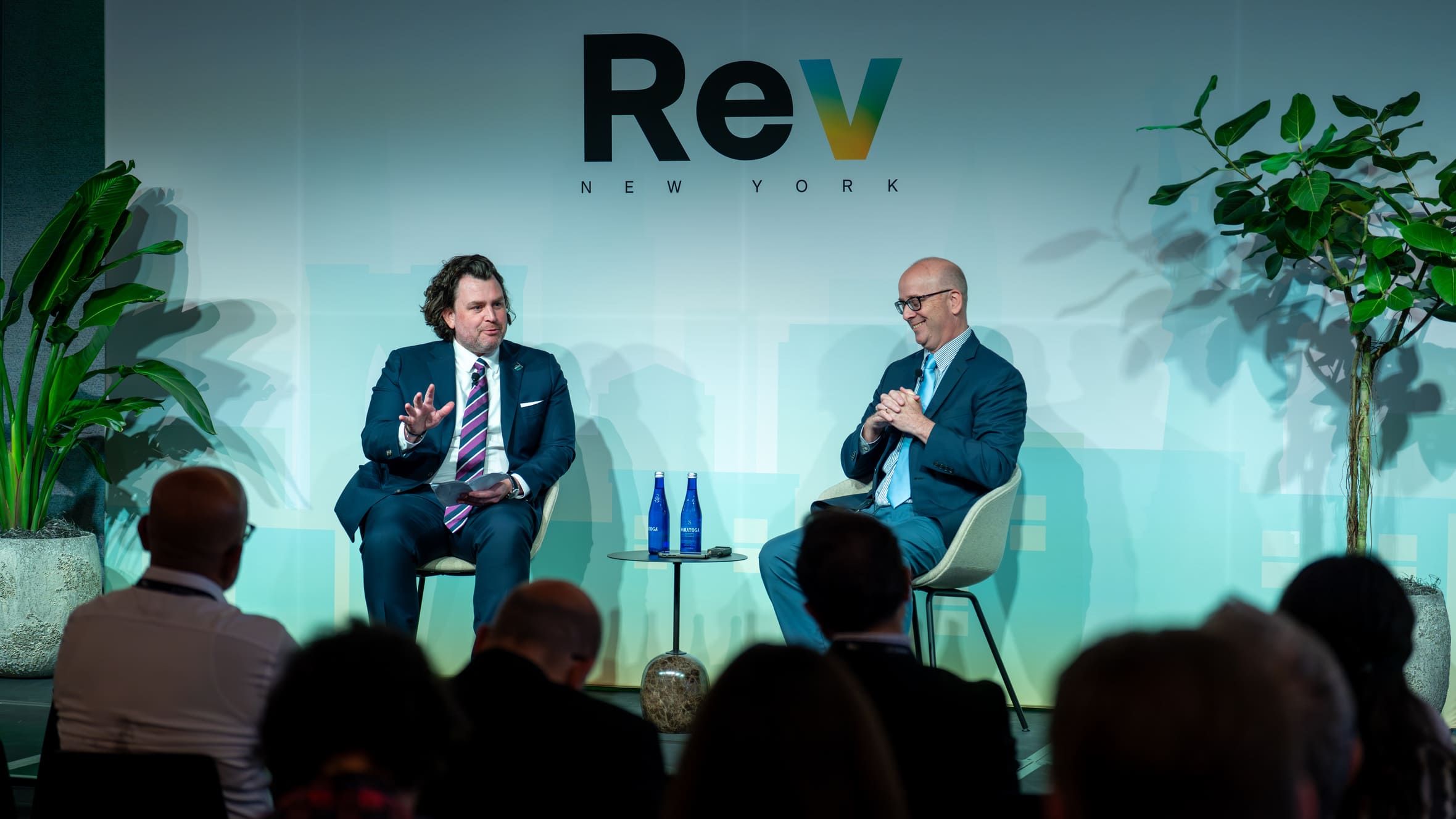 Two men in suits having a discussion on stage with Rev New York logo behind them and an audience in front.