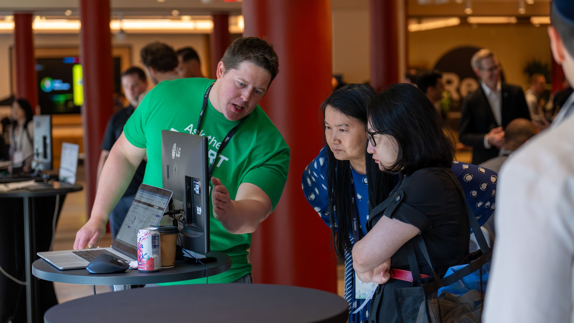 Man in green shirt showing a computer screen to two women in a busy indoor event space.