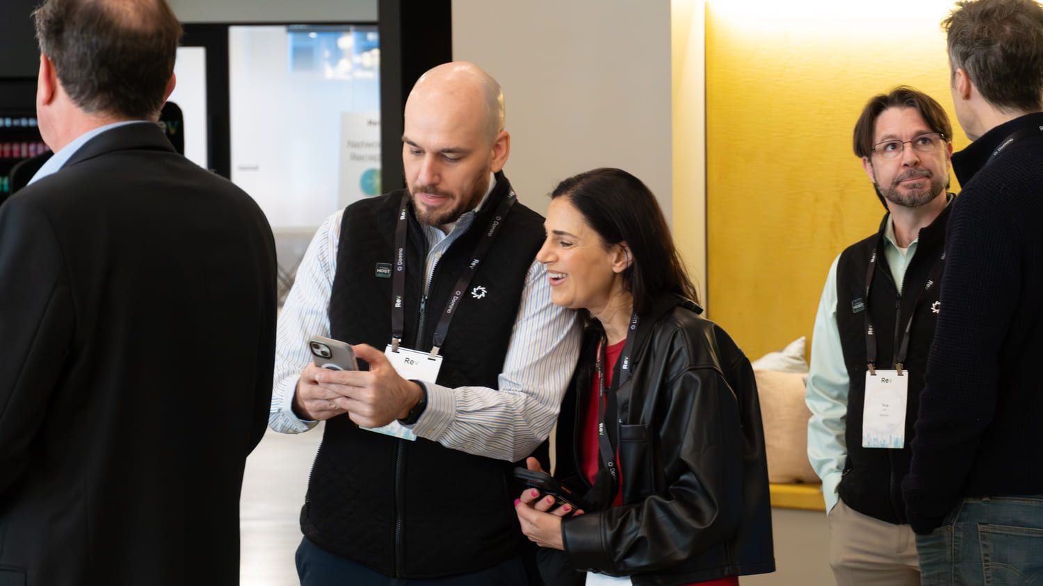 Two men and a woman wearing conference badges chatting and looking at phones in a modern indoor space.