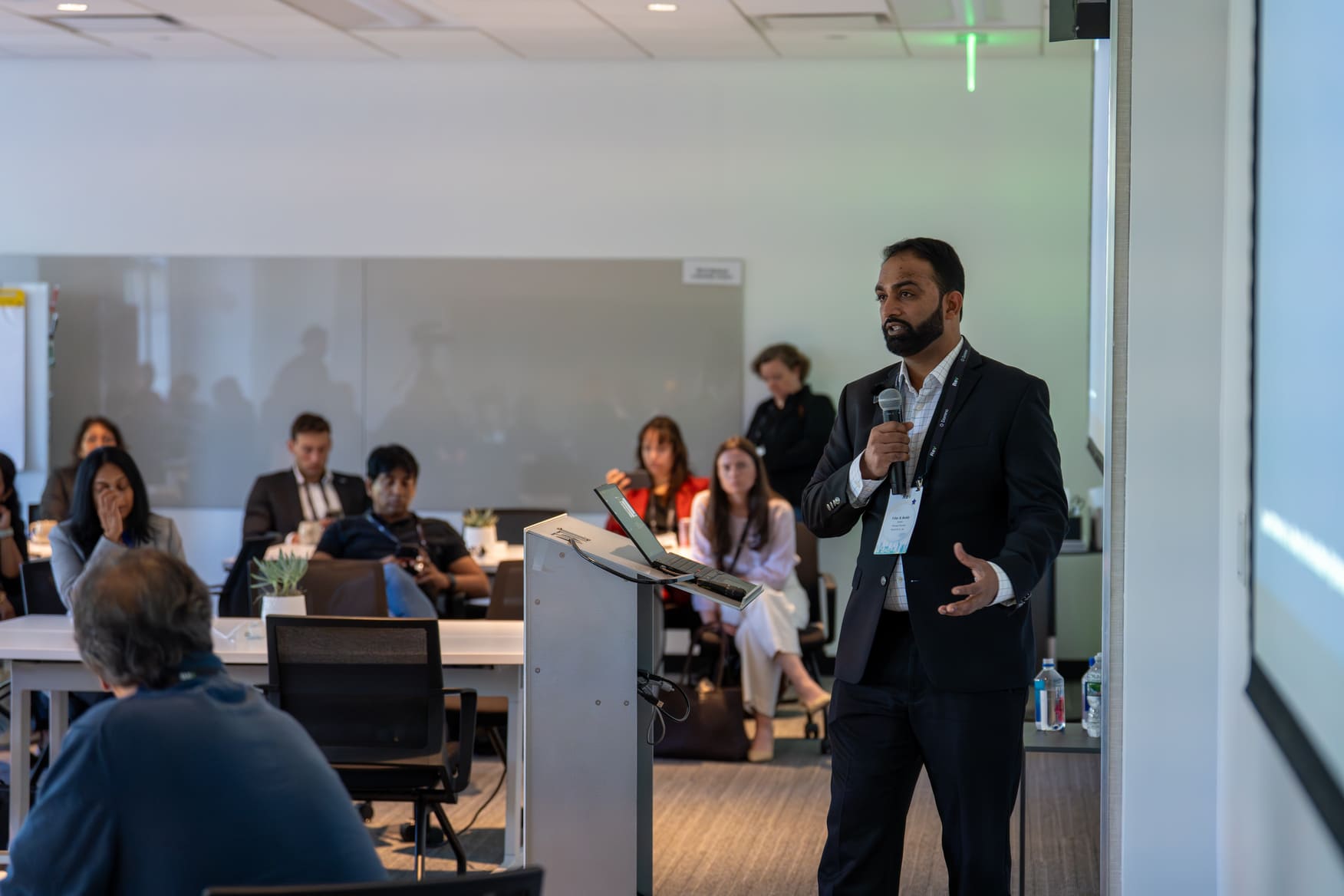 Man in a suit holding a microphone presenting to seated audience in a conference room.