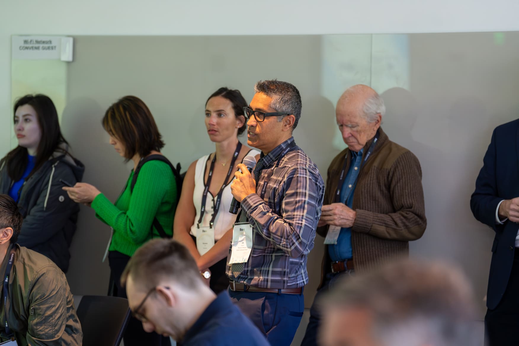 Man with glasses speaks into microphone as others stand or sit in a conference room.
