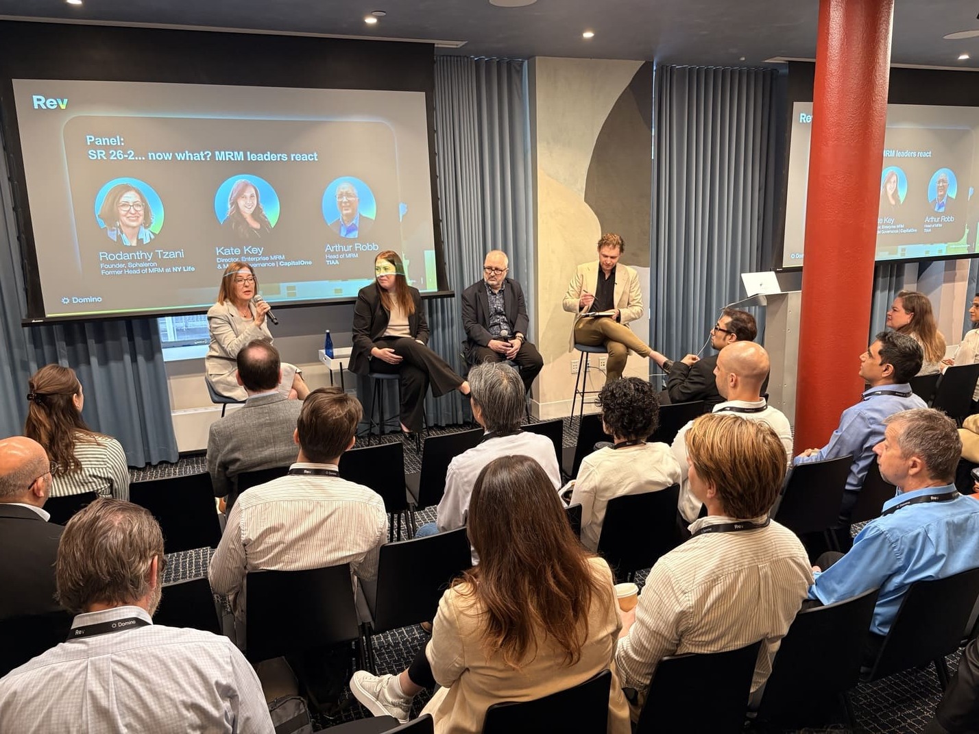 Panel discussion with four speakers and an audience in a conference room with presentation screens.
