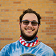 Smiling person with glasses and a colorful neck gaiter against a brick wall background.