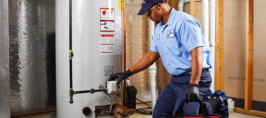 Technician in blue uniform and gloves inspecting a residential water heater in a basement.