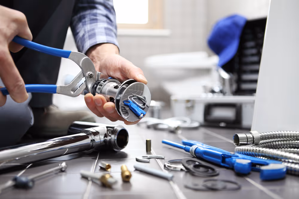 Person using pliers to fix a chrome plumbing pipe with various plumbing tools and parts on a dark tiled surface.