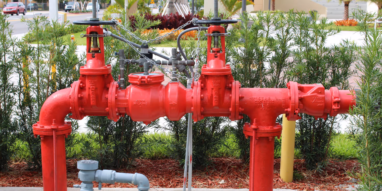 Red industrial backflow valve assembly with pipes and control chains installed outdoors with greenery in the background.