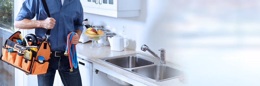 Plumber in blue shirt holding an orange tool bag and red and blue hoses beside a double kitchen sink.