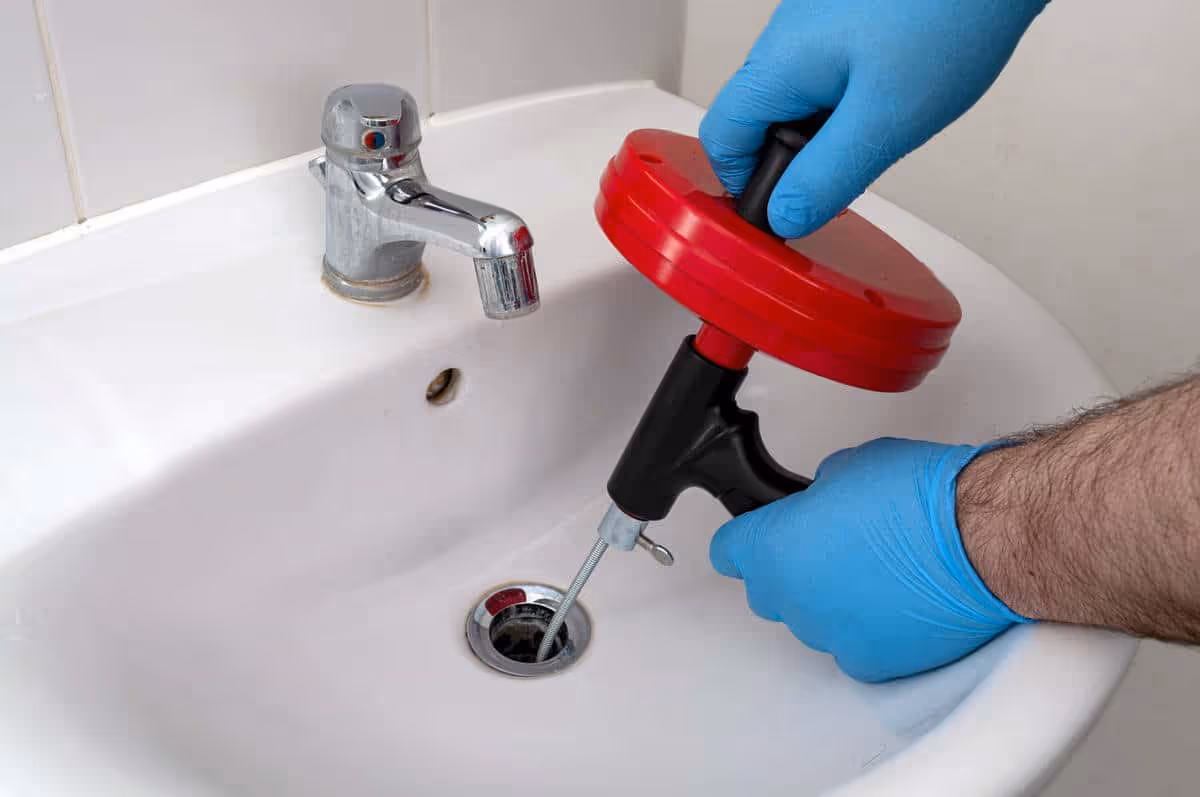 Person wearing blue gloves using a drain snake tool to unclog a sink drain with a chrome faucet.