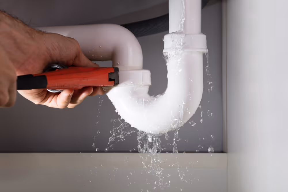 Hand using a red pipe wrench to fix a leaking white U-shaped drain pipe under a sink with water splashing out.