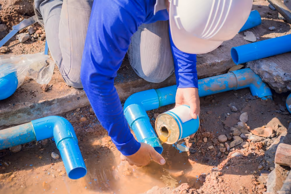 Person in a white hard hat and blue long sleeve shirt holding a water filter near blue PVC pipes in muddy soil.