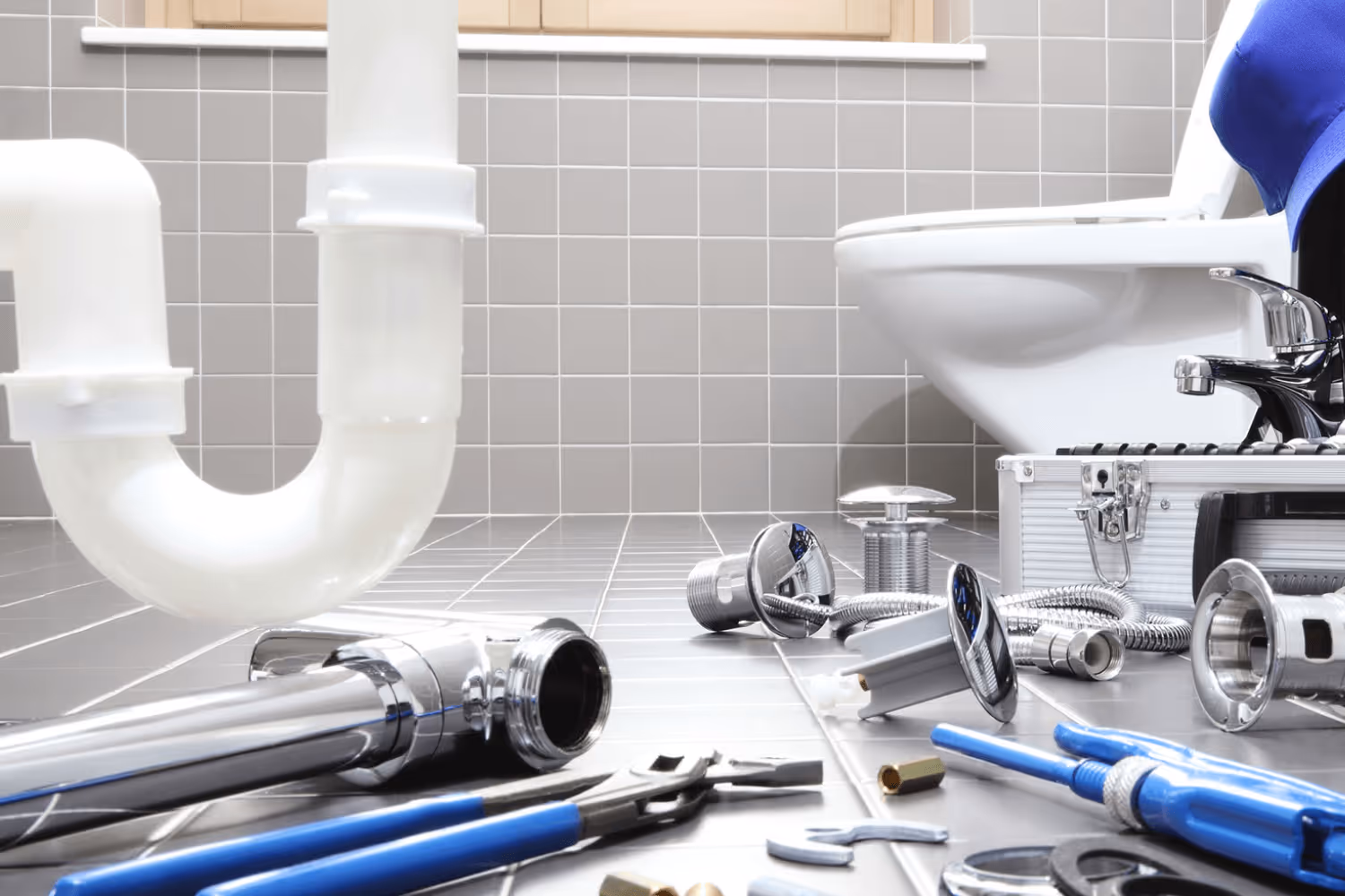 Plumbing tools and parts scattered on a tiled bathroom floor next to a toilet with exposed white pipes.