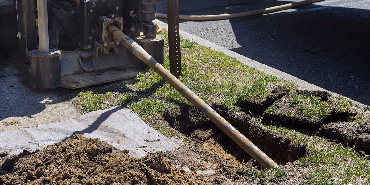 Drilling machine with a long drill bit boring into the ground next to a grassy area and a pile of dirt.