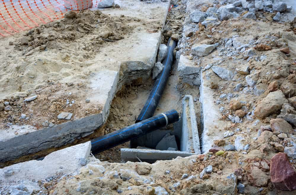 Excavated ground with exposed black sewer pipes and concrete debris surrounded by dirt and rocks.