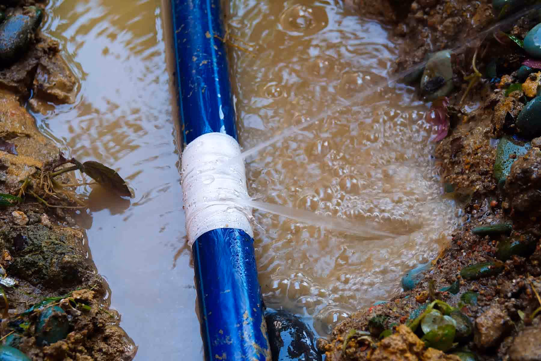 Blue water pipe leaking water from a white taped section into muddy ground.