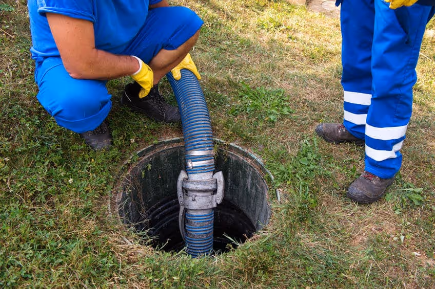 Two workers in blue uniforms with yellow gloves handling a large hose inserted into a grassy sewer opening.