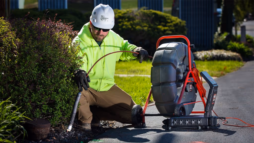 Worker wearing a white hard hat, neon green hoodie, and sunglasses operating a drain cleaning machine outdoors near bushes.