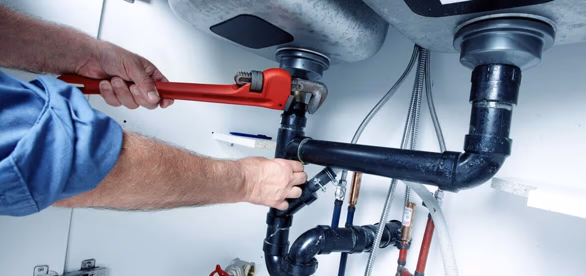 Person using a red pipe wrench to tighten black plumbing pipes under a sink.