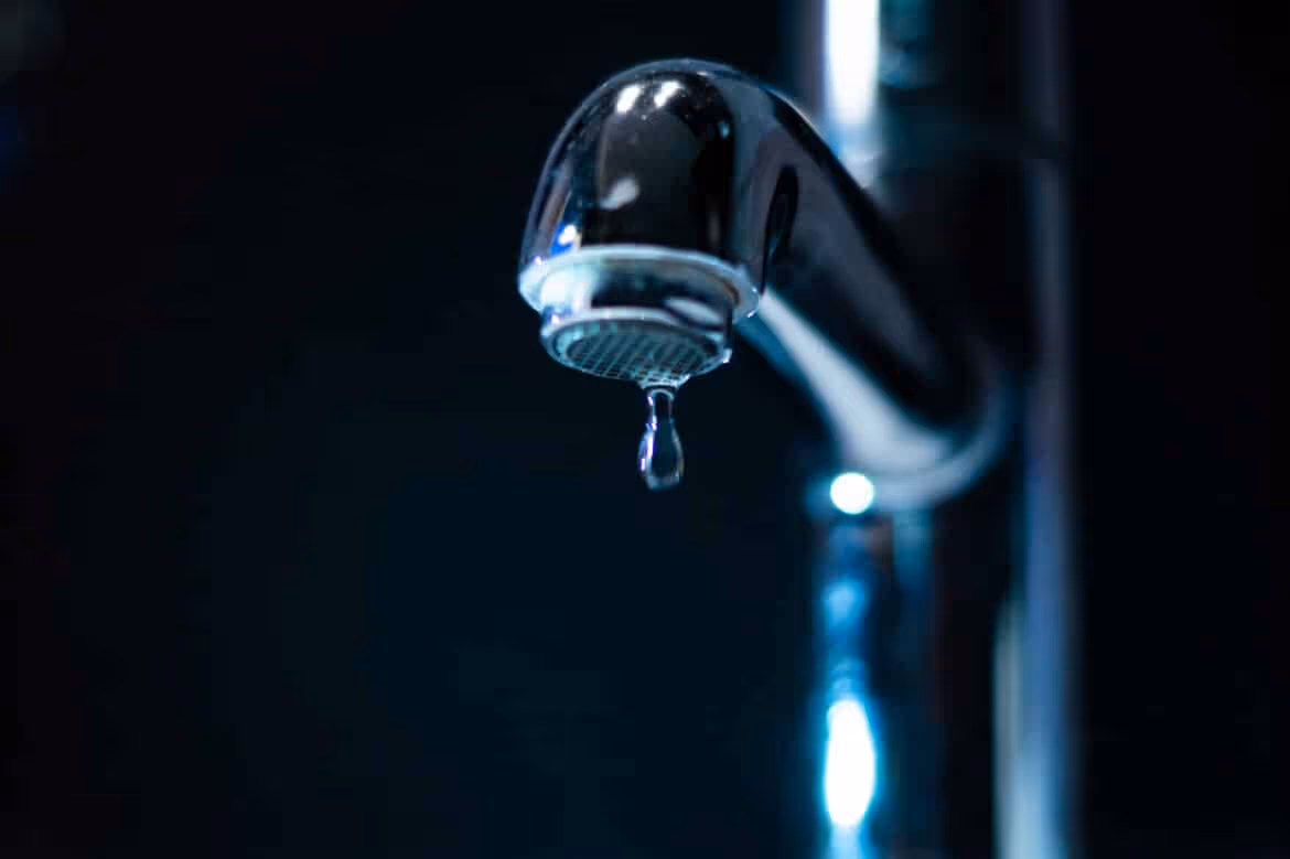 Close-up of a chrome water faucet with a single droplet hanging from the spout against a dark background.
