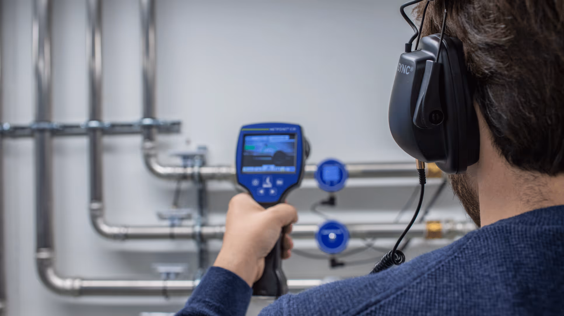 Technician wearing headphones uses a handheld ultrasonic device to inspect pipes for leaks.