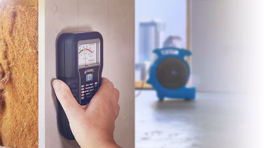 Hand holding a moisture meter against drywall with insulation visible on the left and a blue air blower in the background indoors.
