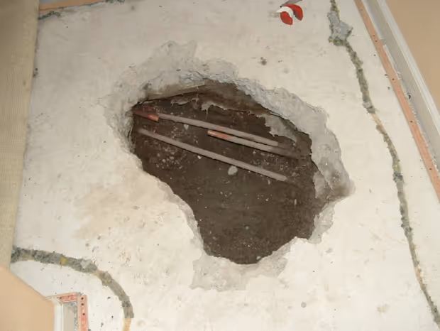 Large irregular hole in concrete floor exposing three horizontal pipes underneath.