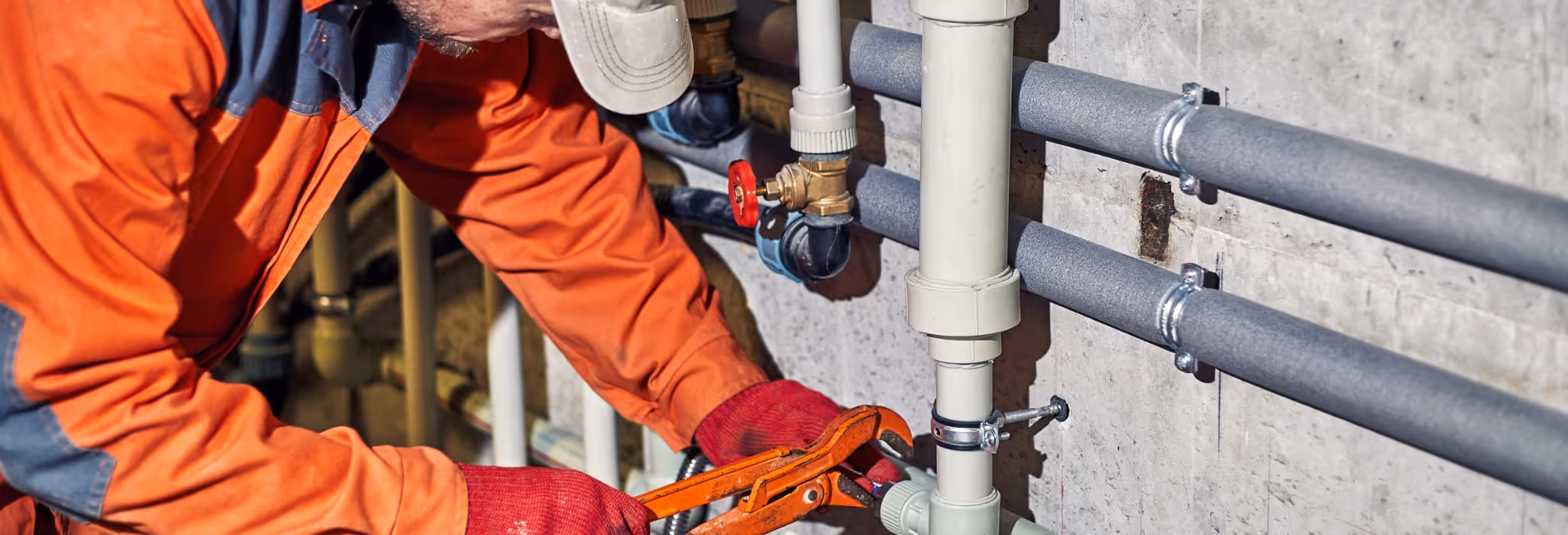 Plumber in orange coveralls and red gloves tightening a pipe fitting with a wrench on wall-mounted plumbing pipes.