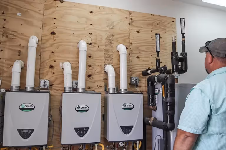 Man in a blue shirt inspecting a row of four A.O. Smith tankless water heaters mounted on a wooden wall.
