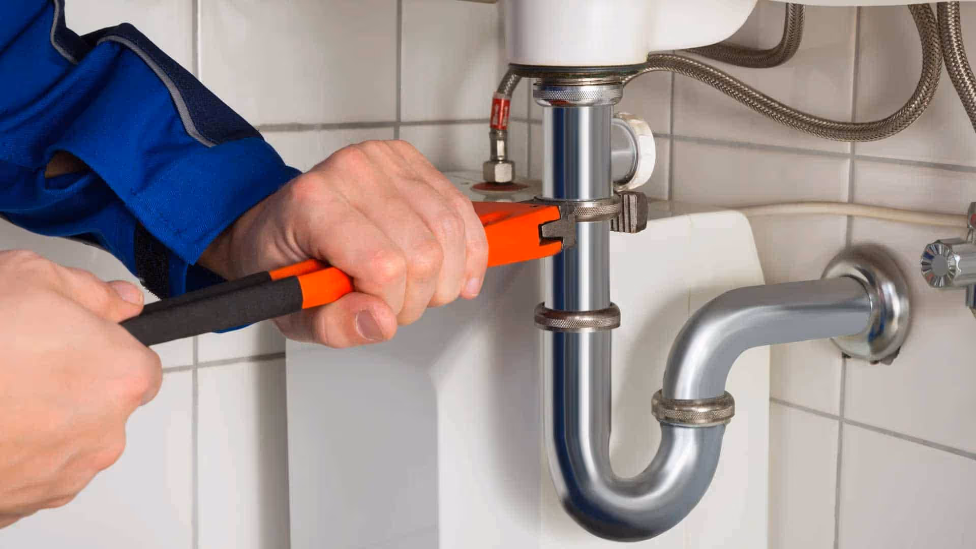 Hands using a red pipe wrench to tighten a chrome pipe under a sink with white tiled wall background.