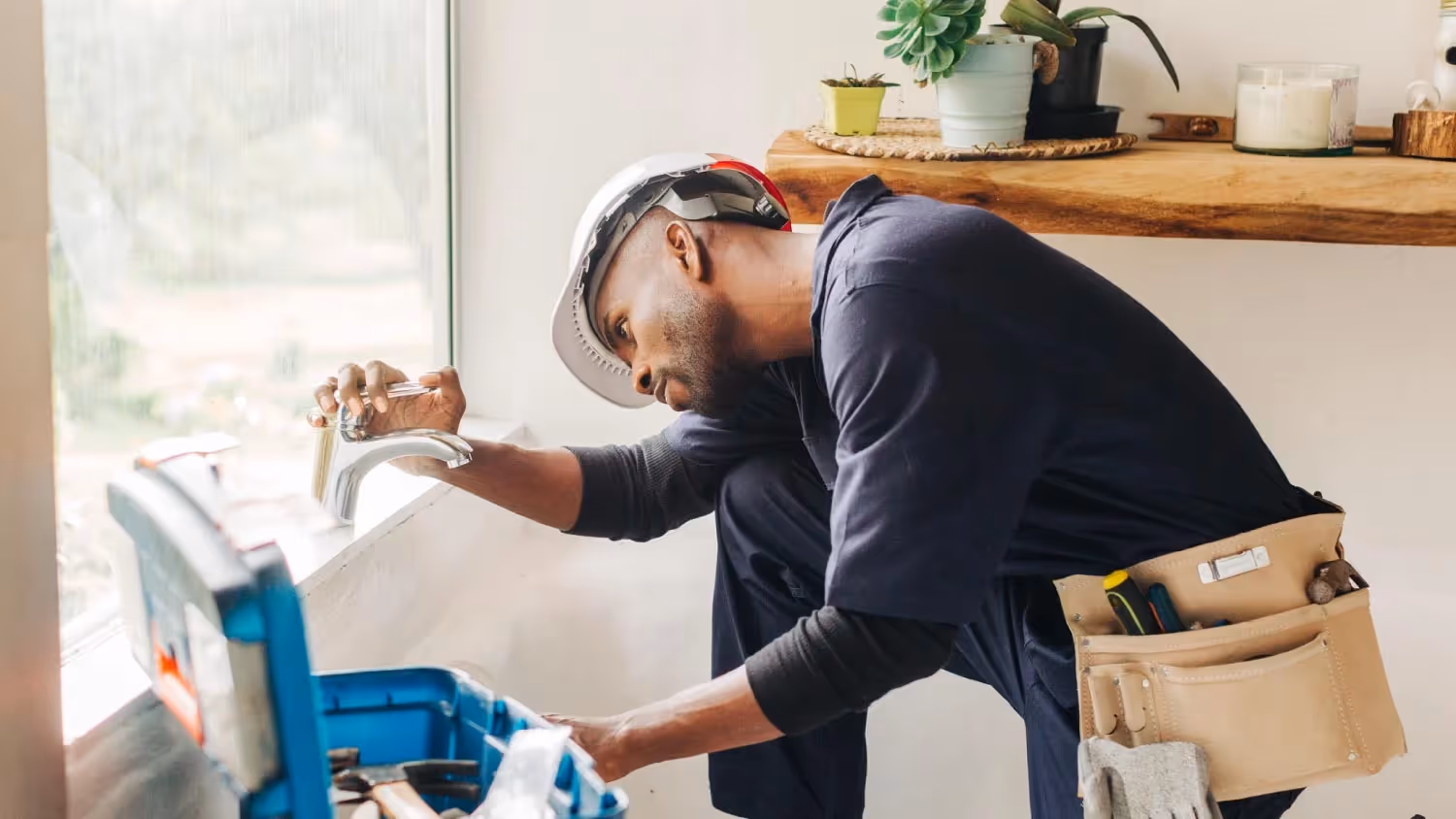 Worker wearing a white helmet and tool belt fixing a faucet next to a blue toolbox under a wooden shelf with potted plants.