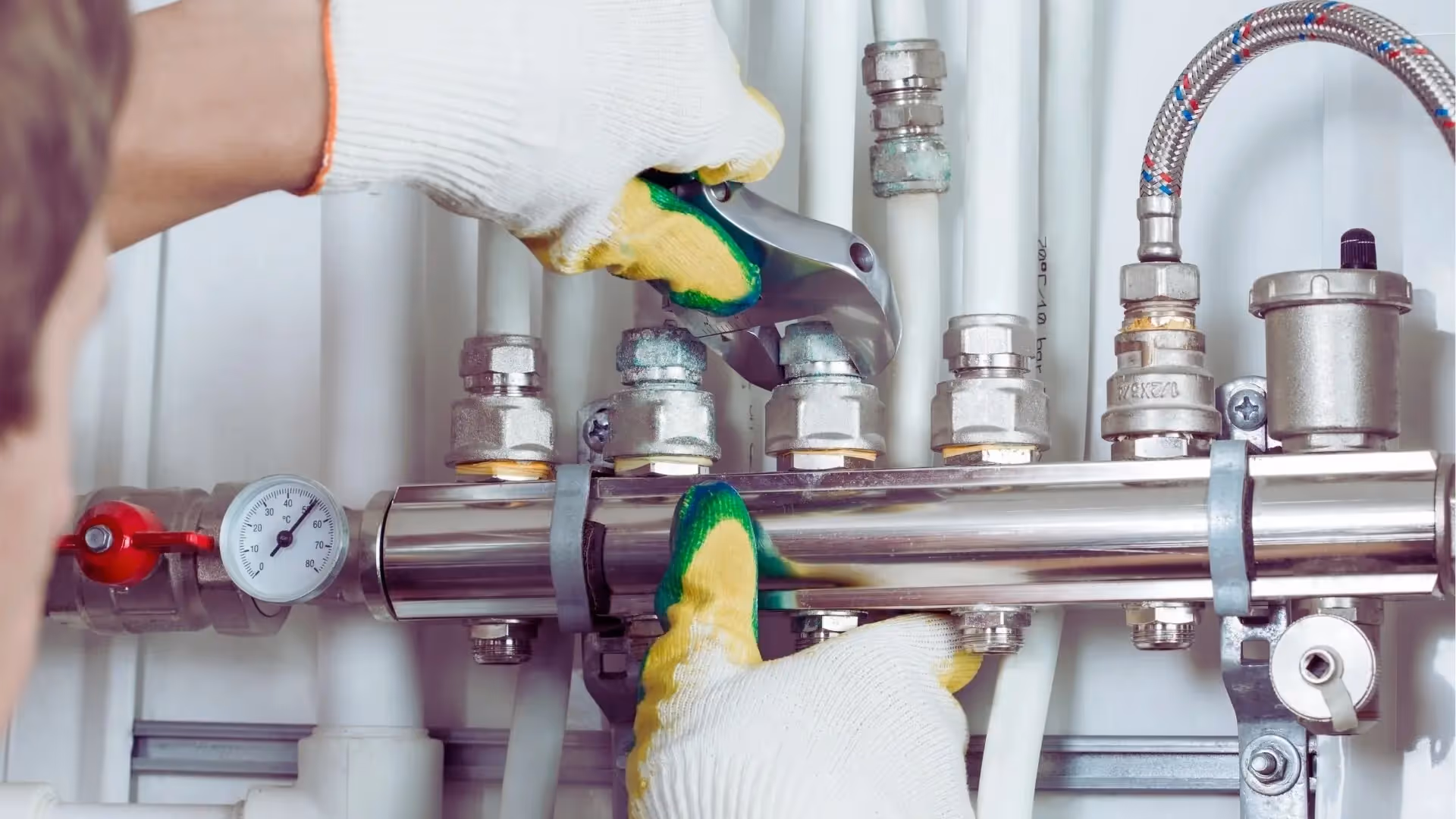 Person wearing gloves using a wrench to tighten a valve on a shiny metal plumbing manifold with connected pipes and a pressure gauge.