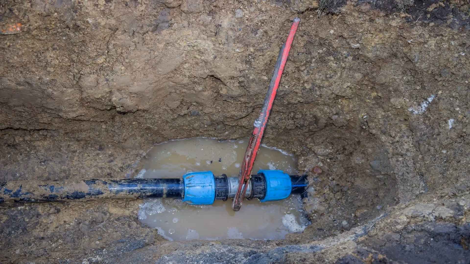 Blue plastic pipe fittings connected underground with a red pipe wrench tightening a joint surrounded by muddy water and soil.