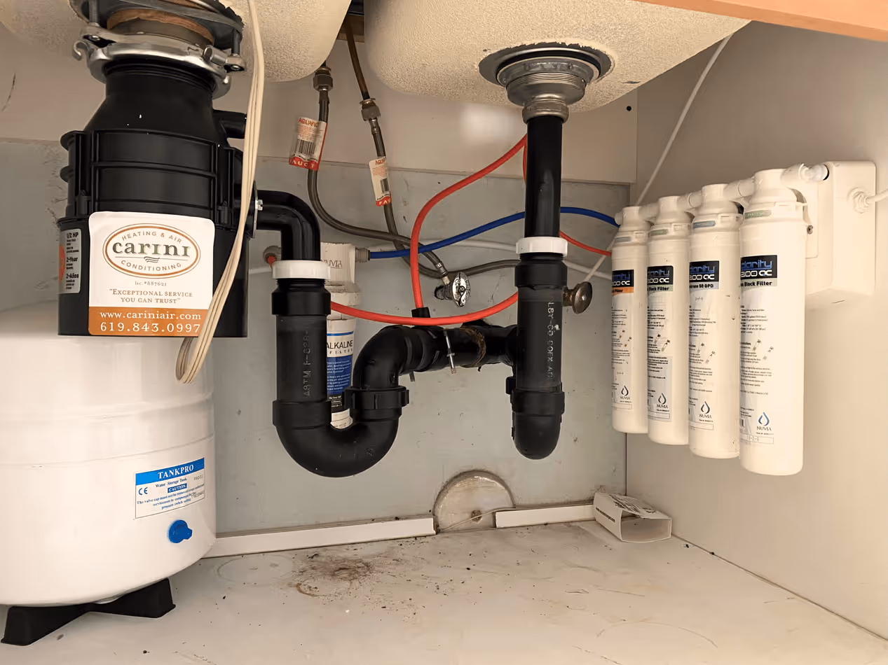 Water filtration system installed under a sink with black plumbing pipes and five white filter cartridges attached to the right interior wall.