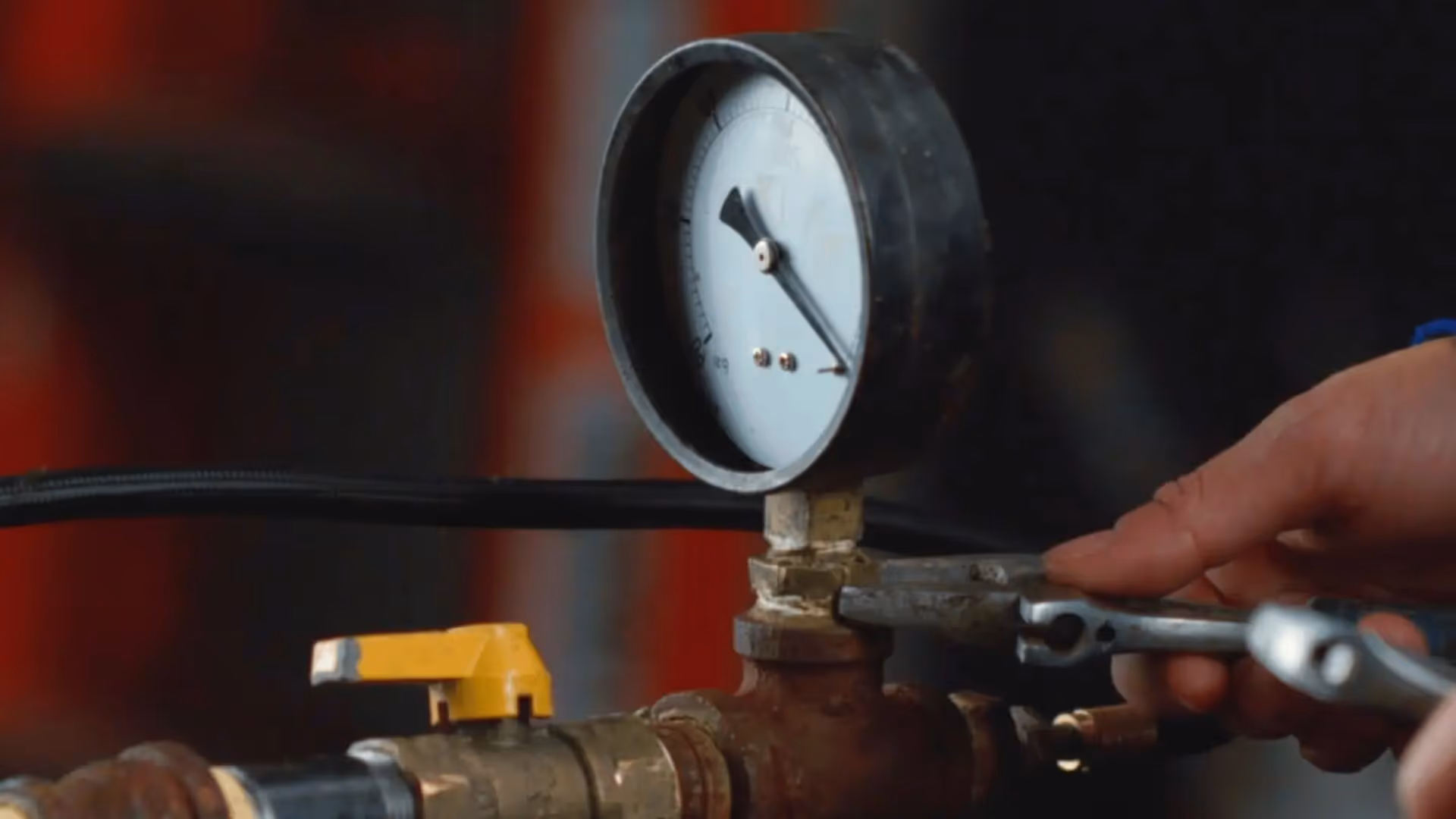 Hand using a wrench to adjust a pressure gauge on a pipe with a yellow valve handle.