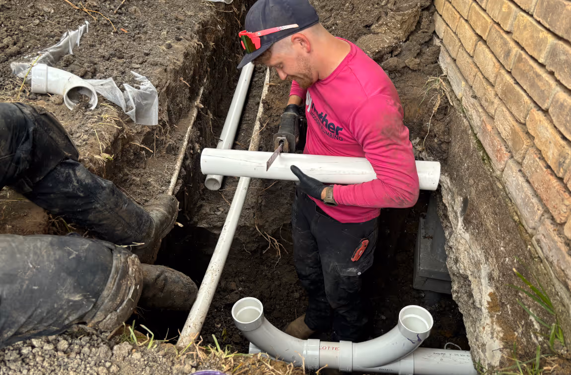 A worker in a pink shirt cutting a white PVC pipe inside a dirt trench next to a brick wall.