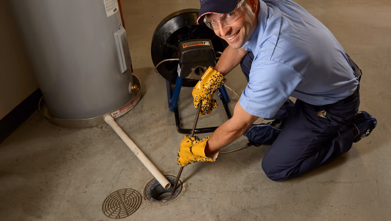 Smiling plumber wearing gloves and safety glasses using a drain snake to unclog a basement floor drain near a water heater.