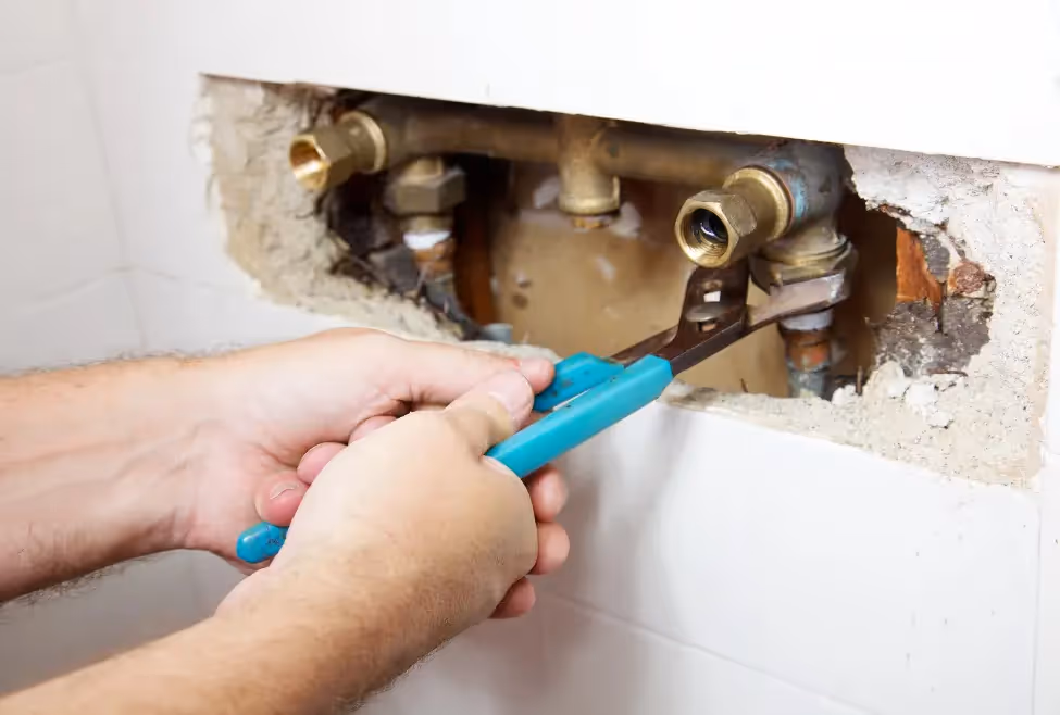 Person using a blue-handled wrench to tighten plumbing fittings inside a wall cavity.