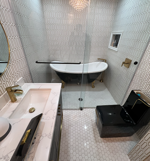 Modern bathroom with a black clawfoot bathtub with gold feet behind a glass shower enclosure, a black toilet, white hexagonal floor tiles, and a marble countertop with a gold faucet.